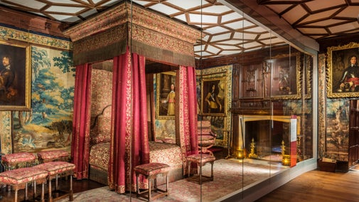 A room with a large framed ornate bed in the centre. The bed has red and gold covers and red curtains hanging at the corners. There is a fireplace to the right, stools to the left and at the foot of the bed and a tapestry on the wall to the left of the bed. There are also portraits hanging on the walls around the room, and the ceiling is white with a pattern created in wood.